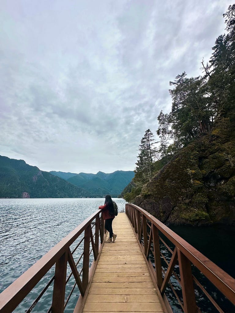 A woman stands on a wooden bridge over a tranquil lake surrounded by forested mountains.