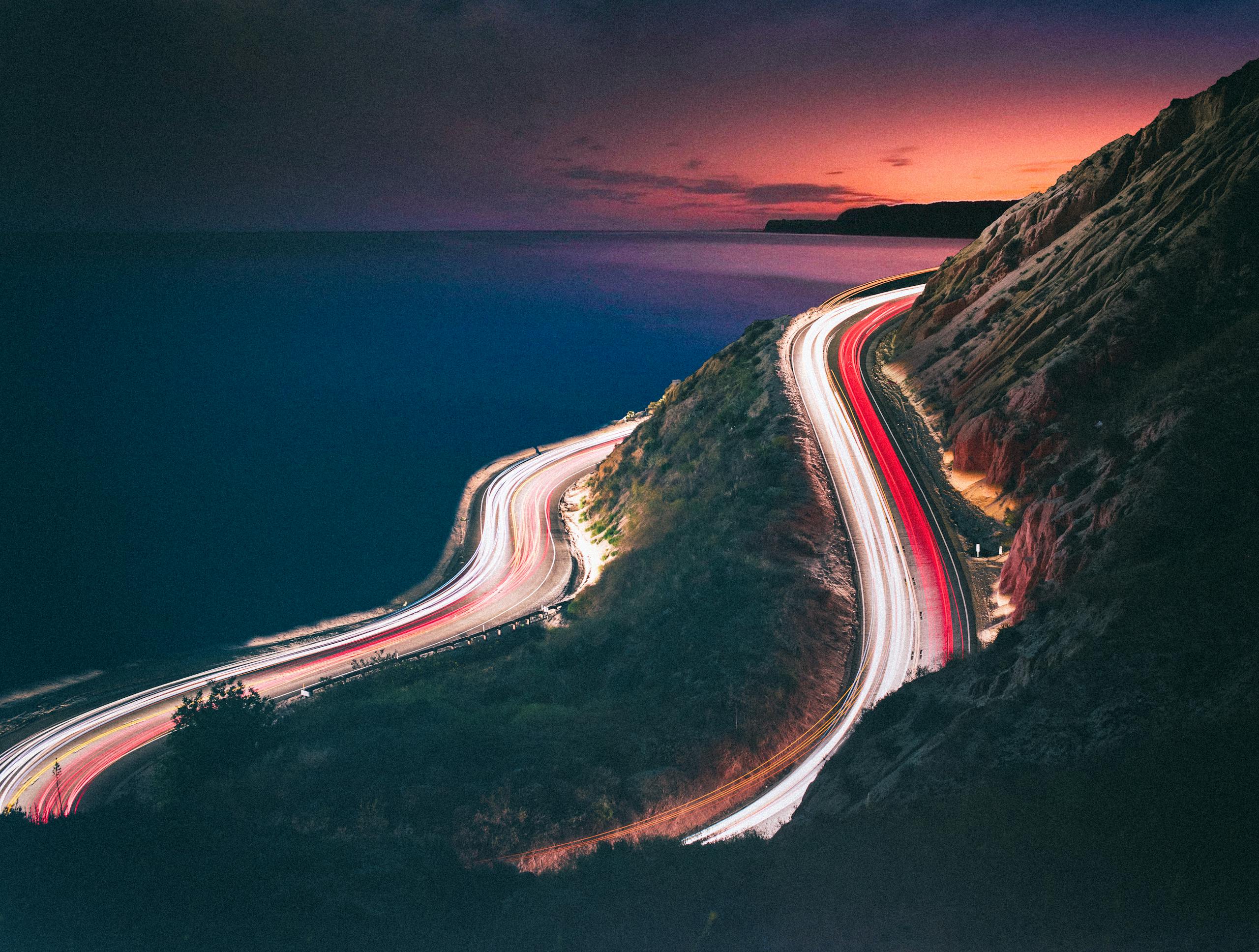 Long exposure night shot of winding coastal road with light trails and ocean view in California.